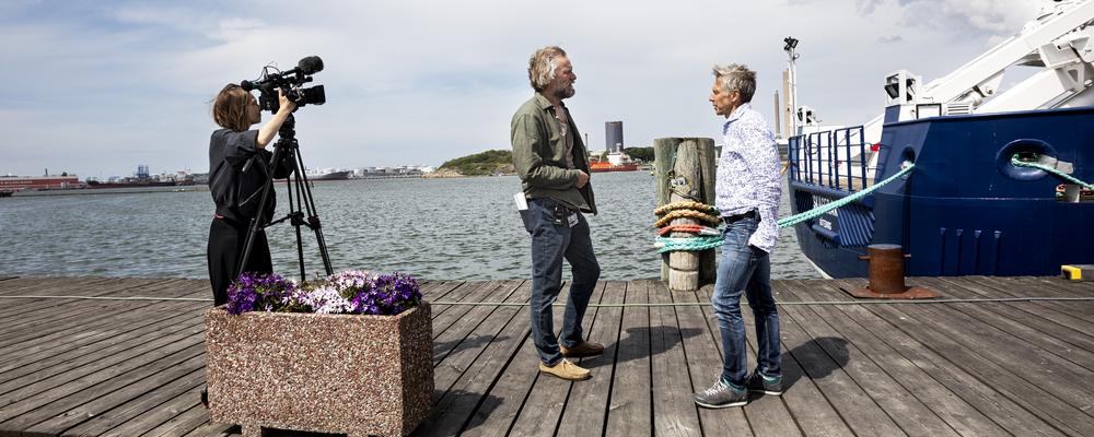 kvinna som lyssnar i hörlurar, man som fotograferar och en hög med tidningar